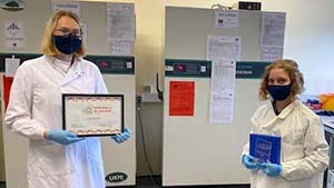 BiobankoftheYear2020_WCB Two people stand at a distance in a lab facing the camera, both wearing face masks. One holds the Biobank of the Year Award certificate while the other holds the tropy